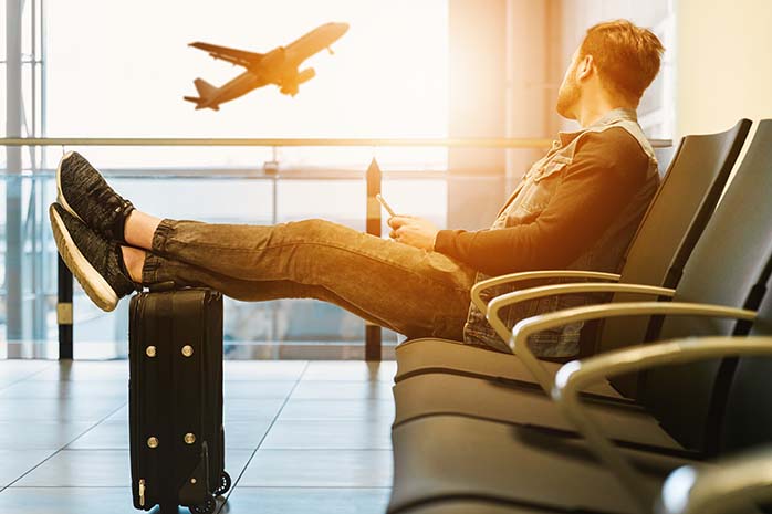 Mann sitzt mit seinem Telefon am Flughafen mit den Füssen auf seinem Koffer. Er guckt auf einen Flugzeug.