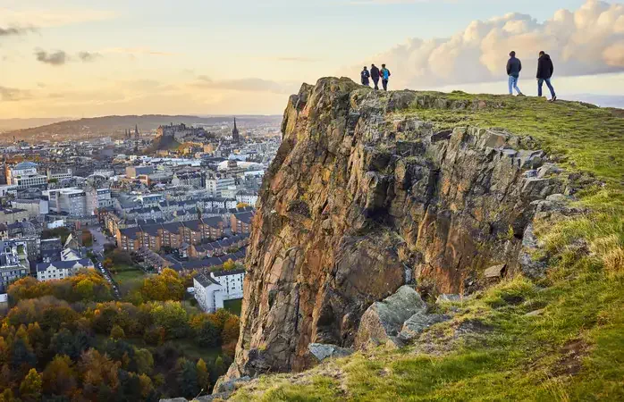 Group of people walking along cliff edge looking at city views. Edinburgh Castle in the distance